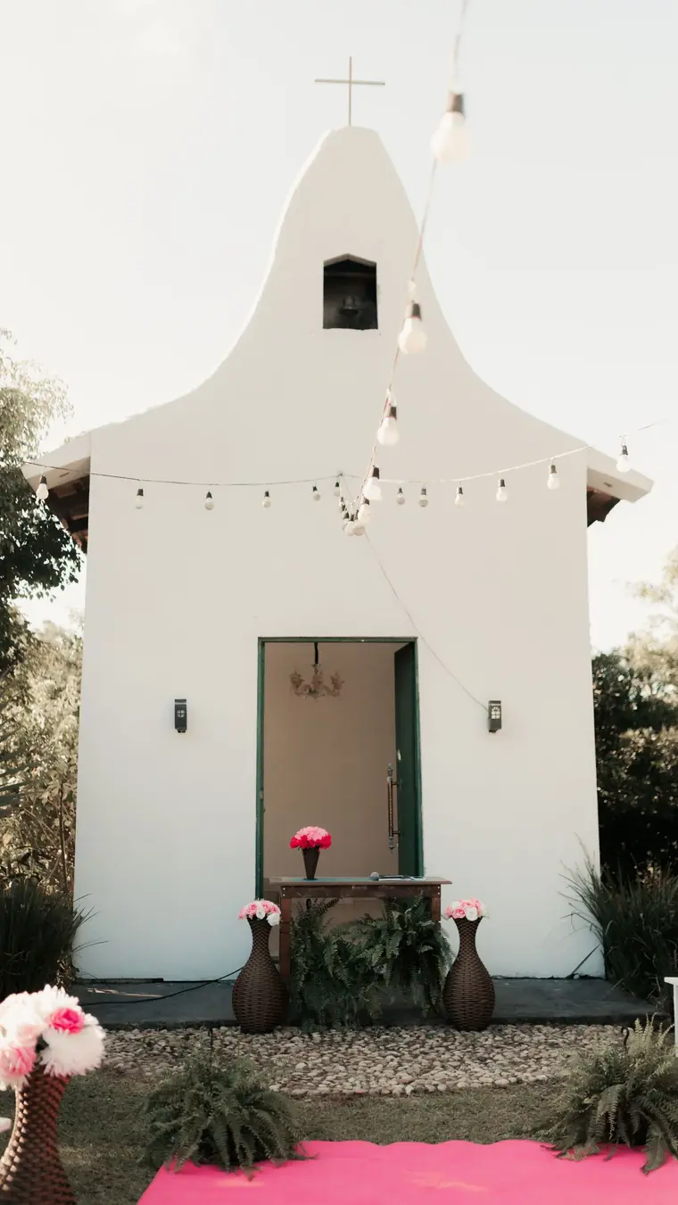 A small white chapel with a curved gable and a bell, festoon lights overhead and a pink carpet leading to pink flowers