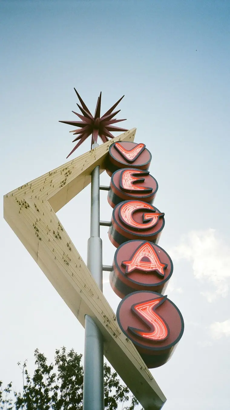 A vintage vertical sign spelling VEGAS in red letters beneath a starburst, seen from below against a pale sky