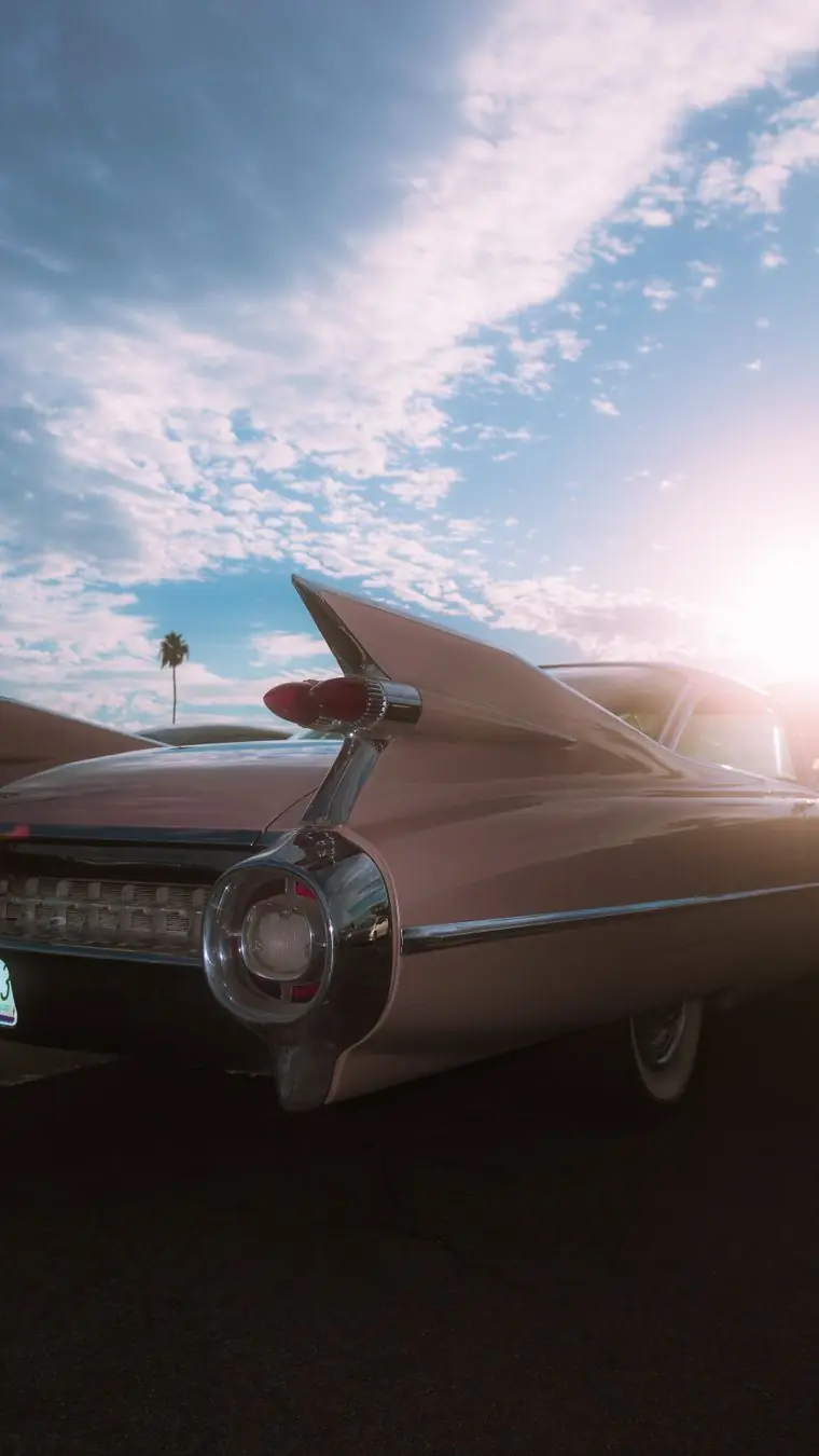 The tail fins of a pink vintage Cadillac against a pale blue sky, sun flaring behind a palm